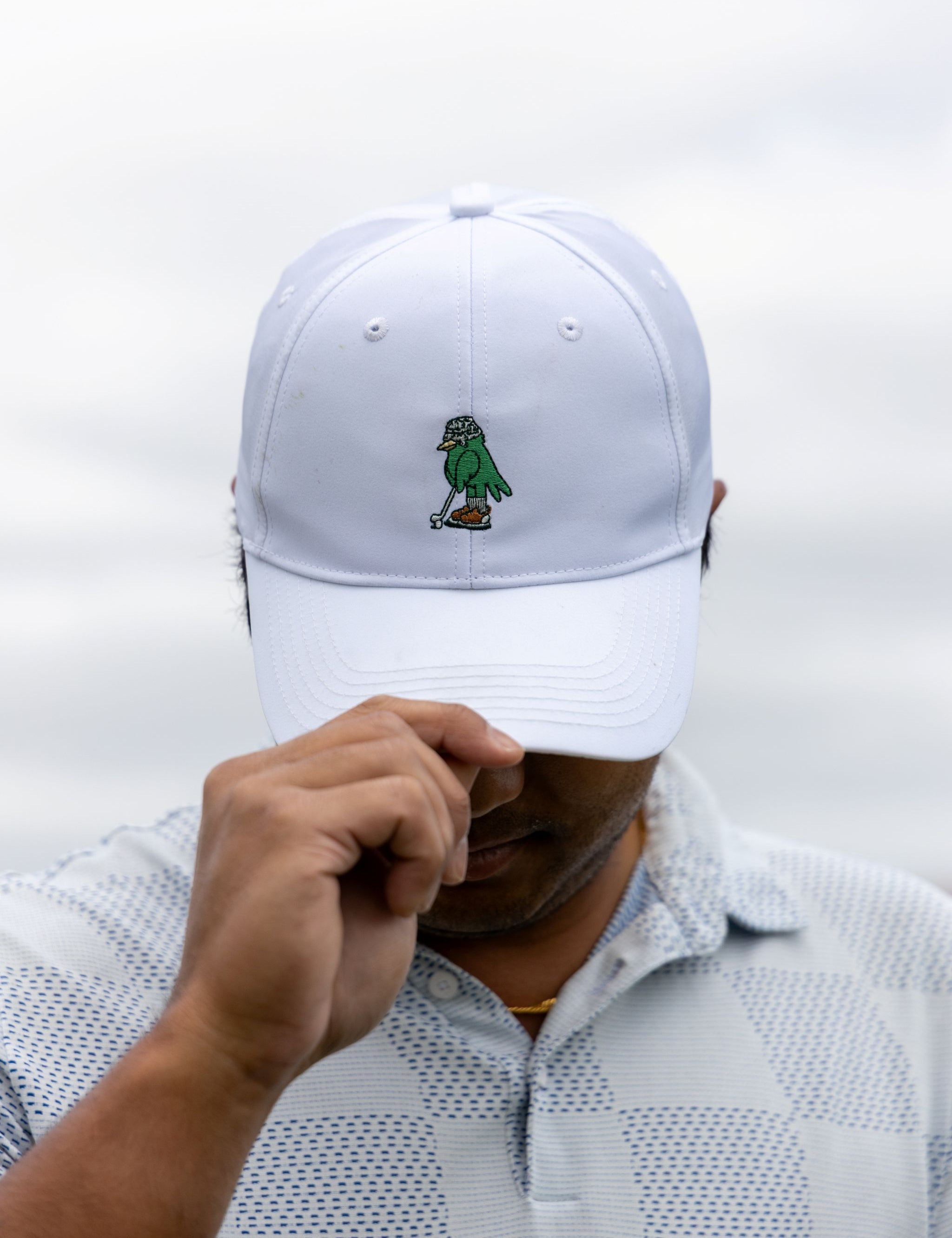 A man wearing a white baseball cap with a small embroidered green bird logo on the front, holding the brim over his eyes. He is wearing a white and gray patterned polo shirt against a soft, overcast sky background.