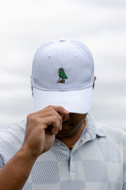 A man wearing a white baseball cap with a small embroidered green bird logo on the front, holding the brim over his eyes. He is wearing a white and gray patterned polo shirt against a soft, overcast sky background.
