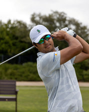A male golfer in a white patterned polo shirt, white cap, and mirrored sunglasses swings a golf club on a grassy course with trees in the background.