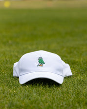 A white baseball cap with a small embroidered green bird logo resting on a green grass field.