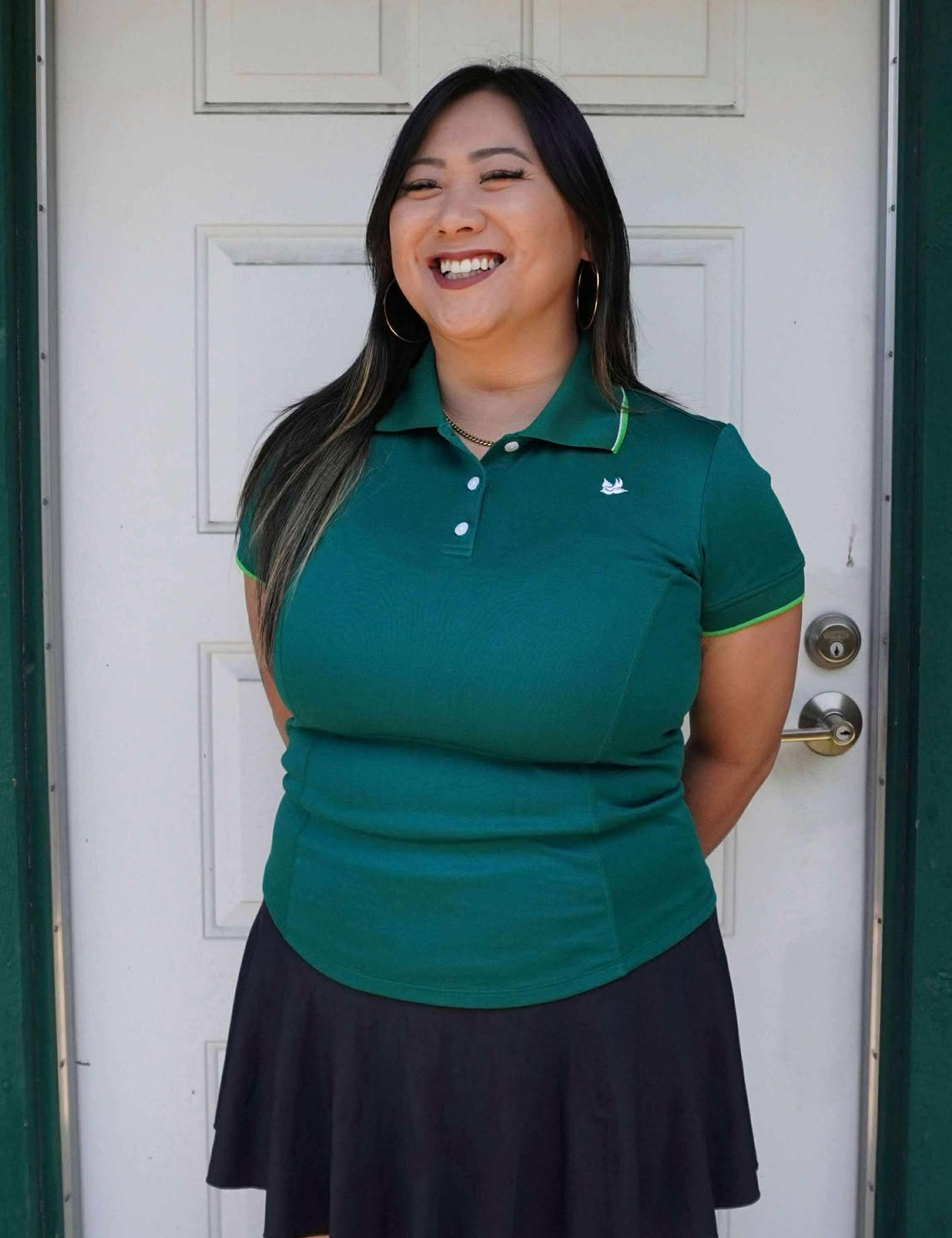 Woman wearing a green polo shirt and black skirt standing in front of a white door.